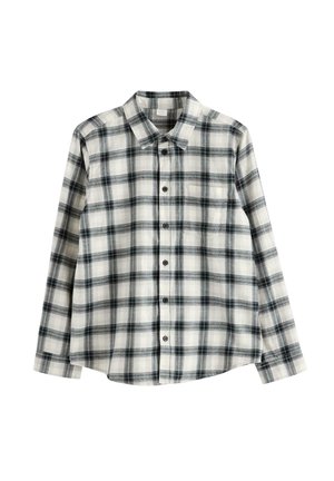Nastolatek w brązowej czapce, czarno-białej kratowanej koszuli na białym t-shircie, czarnych jeansach i białych trampkach, stojący na jednolitym tle.