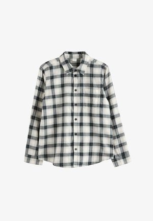 Teenager iført en brun beanie, sort-hvid ternet skjorte over en hvid t-shirt, sorte jeans og hvide sneakers, der står mod en ensfarvet baggrund.