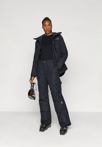 Black waterproof jacket with a hood and zippered pockets paired with matching cargo pants. Model holds ski goggles, wearing black boots.