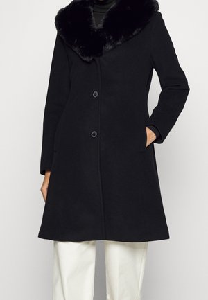 Abrigo negro hasta media pierna con cuello de piel negro y cierre de botones, llevado sobre una blusa negra y pantalones blancos, con las manos en los bolsillos laterales.