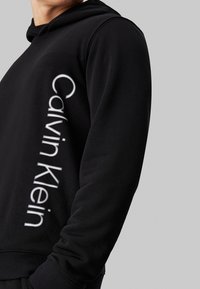 Felpa nera con cappuccio realizzata in materiale morbido, caratterizzata da un evidente logo verticale bianco "Calvin Klein" lungo la manica sinistra. Polsini elasticizzati.