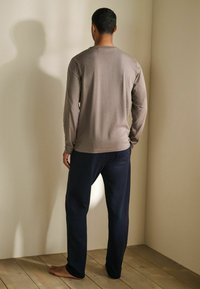 Camicia marrone a maniche lunghe e pantaloni da pigiama blu navy. Materiale in cotone morbido, vestibilità rilassata, cuciture minime e design semplice. A piedi nudi su un pavimento di legno.