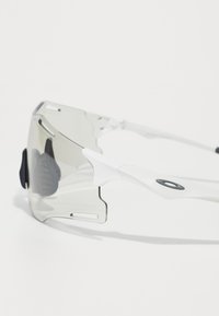 Weiße Sportsonnenbrille mit gebogenen, transparenten grauen Gläsern, die ein einzigartiges Design mit offenen Seiten und strukturierten schwarzen Gummi-Akzenten aufweist.
