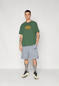 Žalia medvilninė marškinėliai su geltonu logotipu, derinami su pilkomis šortais. Modelis dėvi pilkas kojines ir baltus sportbačius su juodais akcentais.