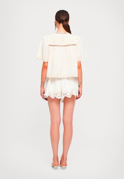 Woman with dark hair in a low ponytail wearing a loose white embroidered blouse and matching lace-trimmed shorts, standing with back to camera.