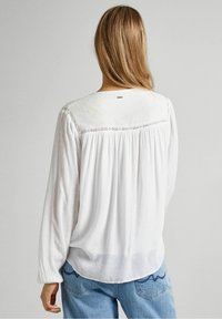 Donna con lunghi capelli biondi che indossa una blusa bianca a maniche lunghe con dettagli in pizzo, abbinata a jeans azzurri, rivolta di spalle rispetto alla fotocamera.
