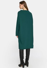 Kobieta ubrana w luźną, długą, ciemnozieloną sukienkę, w połączeniu z czarnymi, wiązanymi wysokimi botkami, widziana od tyłu na białym tle.