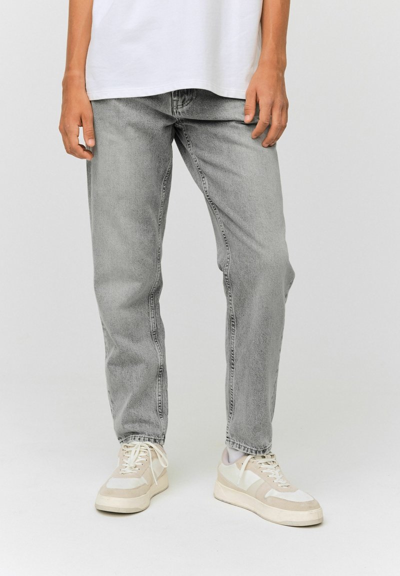 Personne portant un jean gris clair, des baskets blanches et beiges, et une chemise blanche avec les mains détendues le long du corps sur un fond uni.