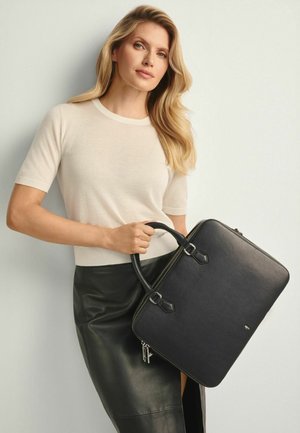 Kobieta o długich blond włosach, ubrana w kremową bluzkę z krótkim rękawem i czarną skórzaną spódnicę, trzymająca dużą czarną skórzaną teczkę.