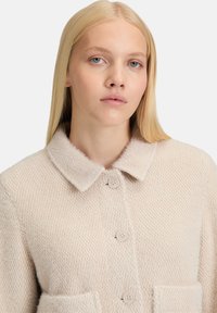 Beige structuurjas met een kraag, vier knopen en twee voorzakken. Het materiaal lijkt zacht en warm.