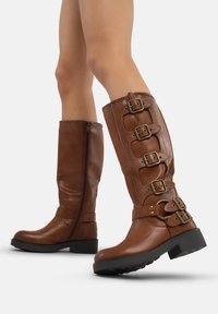 Botas de cuero marrón hasta la rodilla con hebillas, acabado texturizado y cremalleras laterales. Suela gruesa con un ligero tacón.