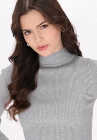 Grauer, gerippter Rollkragenpullover mit schmalem Schnitt. Glatte Textur mit leichtem Stretch. Das Model hat lange, wellige Haare und kleine Ohrringe.
