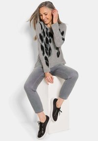 Grauer Pullover mit schwarzen Leopardenflecken, kombiniert mit grauen Hosen und schwarzen Turnschuhen. Model sitzt auf einem weißen Block. Innenaufnahme.