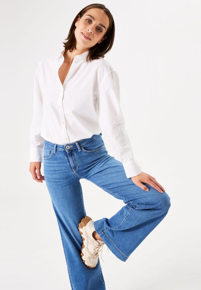 Witte button-up blouse met lange mouwen, blauwe denim uitlopende jeans en lichtbeige sneakers. De stof lijkt soepel en heeft een relaxte pasvorm.