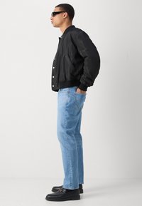 Homme portant une veste bomber noire, un jean bleu clair, des mocassins noirs et des lunettes de soleil, se tenant de profil les mains dans les poches devant un fond uni.