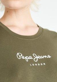 Olive spalvos medvilnės marškinėliai su apvaliu kaklu ir baltu "Pepe Jeans London" logotipu, atspausdintu kersiu šriftu per krūtinę.