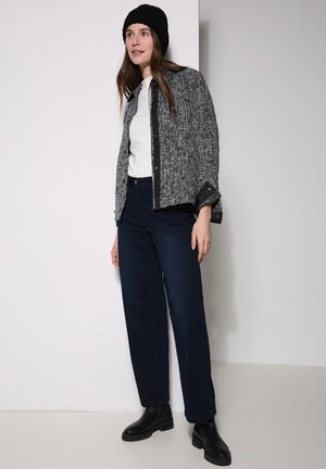 Grau und schwarz strukturierte Jacke über einem weißen Top, kombiniert mit dunkelblauen weiten Hosen und schwarzen Knöchelstiefeln, vor einem neutralen Hintergrund.
