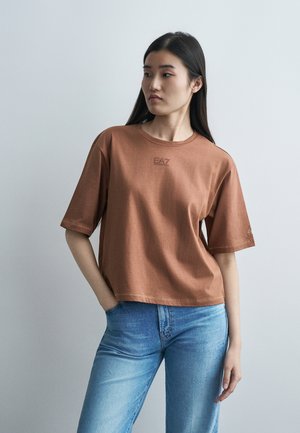 Mladá žena s dlhými tmavými vlasmi má na sebe hnedé tričko EA7 Emporio Armani a svetlomodré džínsy, stojí pred jednoduchým svetlosivým pozadím.