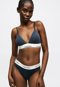 Femme portant un bralette Calvin Klein bleu foncé et un slip assorti avec des bandes élastiques blanches et le logo, debout devant un fond clair uni.