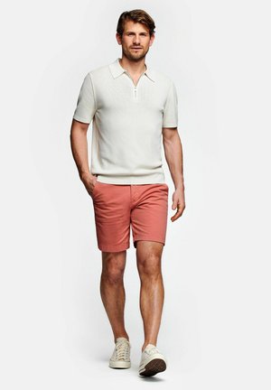Uomo che cammina indossando una polo bianca a maniche corte, pantaloncini color salmone e sneakers crema su uno sfondo bianco semplice.