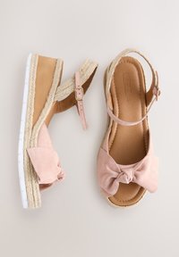 Roze sleehaksandalen met enkelbandjes, strikdetail aan de voorkant, met jute bedekte zolen en witte geribbelde rubberen buitenzolen tegen een neutrale achtergrond.