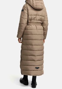 Abrigo de invierno largo acolchado beige con capucha y cinturón, llevado sobre botas gruesas negras, mostrado desde detrás sobre un fondo blanco.