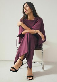 Bordeaux todelt outfit bestående af en løs bluse og tapered bukser, lavet af glat stof. Sorte hælede sandaler med ankelremme.