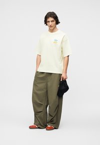 Νεαρός άνδρας με σκούρα μαλλιά που φοράει εκρού μπλούζα, φαρδιά χακί παντελόνια, κόκκινα αθλητικά παπούτσια, κρατώντας ένα μαύρο καπέλο στο δεξί του χέρι.