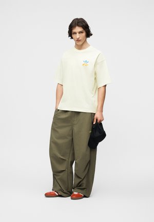 Νεαρός άνδρας με σκούρα μαλλιά που φοράει εκρού μπλούζα, φαρδιά χακί παντελόνια, κόκκινα αθλητικά παπούτσια, κρατώντας ένα μαύρο καπέλο στο δεξί του χέρι.