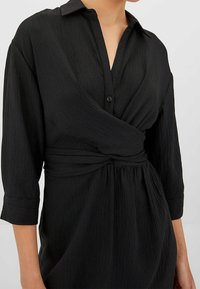 Vestido negro abotonado con tela texturizada, diseño cruzado y mangas tres cuartos. Cuenta con cintura anudada y cuello.
