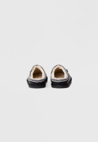 Czarne klapki z miękkim beżowym wnętrzem, zaokrąglonym tyłem, ozdobnym przeszyciem wokół otworu oraz teksturowaną czarną podeszwą.