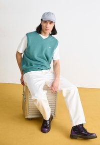 Gilet in maglia turchese sopra una t-shirt bianca, abbinato a pantaloni bianco sporco e stivali nero-viola. Seduto su un contenitore metallico.
