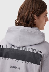 Grijze hoodie met een gestructureerd zwart paneel met ingedeukte letters en accenten. Inclusief een capuchon met trekkoorden en een losse pasvorm.