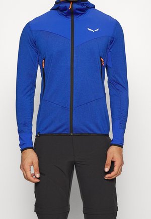 Homme portant une veste à capuche bleue zippée avec des tirettes de fermeture éclair orange et un pantalon noir, debout devant un fond uni.