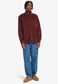 Camisa de pana burdeos con botones, de manga larga y un bolsillo en el pecho, combinada con jeans de denim azul y zapatos de ante marrón.
