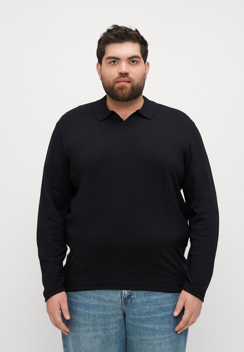 Svart langermet skjorte med V-hals og krage, laget av et mykt materiale, kombinert med lyseblå jeans, stående mot en hvit bakgrunn.