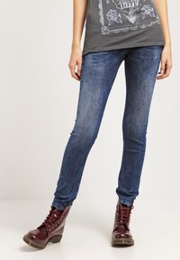 Dark blue skinny jeans made from denim, featuring a smooth texture, snug fit, and subtle fading. Paired with maroon lace-up boots.