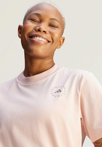 Light pink cotton t-shirt with a round neckline, featuring a circular logo design in gray on the chest. Smooth texture, casual design.