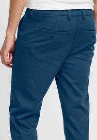 Moški nosi prilagojene temno modre hlače z gumbi na zadnjih žepih in belo srajco, stoji z levo roko sproščeno ob telesu.