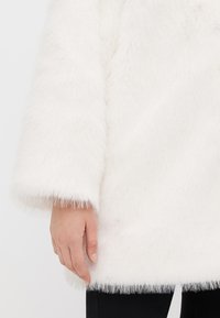 Cappotto in pelliccia sintetica bianca con una texture morbida e soffice e un taglio dritto. Presenta maniche lunghe con bordo in pelliccia ai polsini e all'orlo.