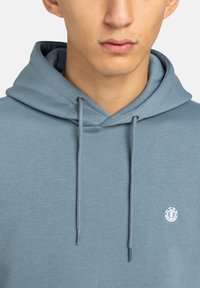 Svetlomodrá mikina s kapucňou a šnúrkou, na ľavej strane hrudníka s malým bielym logom. Mäkky, textúrovaný materiál bez vzorov.