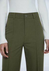 Pantalons ajustés vert olive avec une texture lisse, dotés d'une fermeture à bouton sur le devant, de passants de ceinture et de coutures latérales nettes.