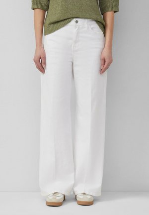 Frau trägt weiße Hose mit weitem Bein, grünen Strickpullover und weiße Turnschuhe, steht vor einem schlichten hellen Hintergrund.