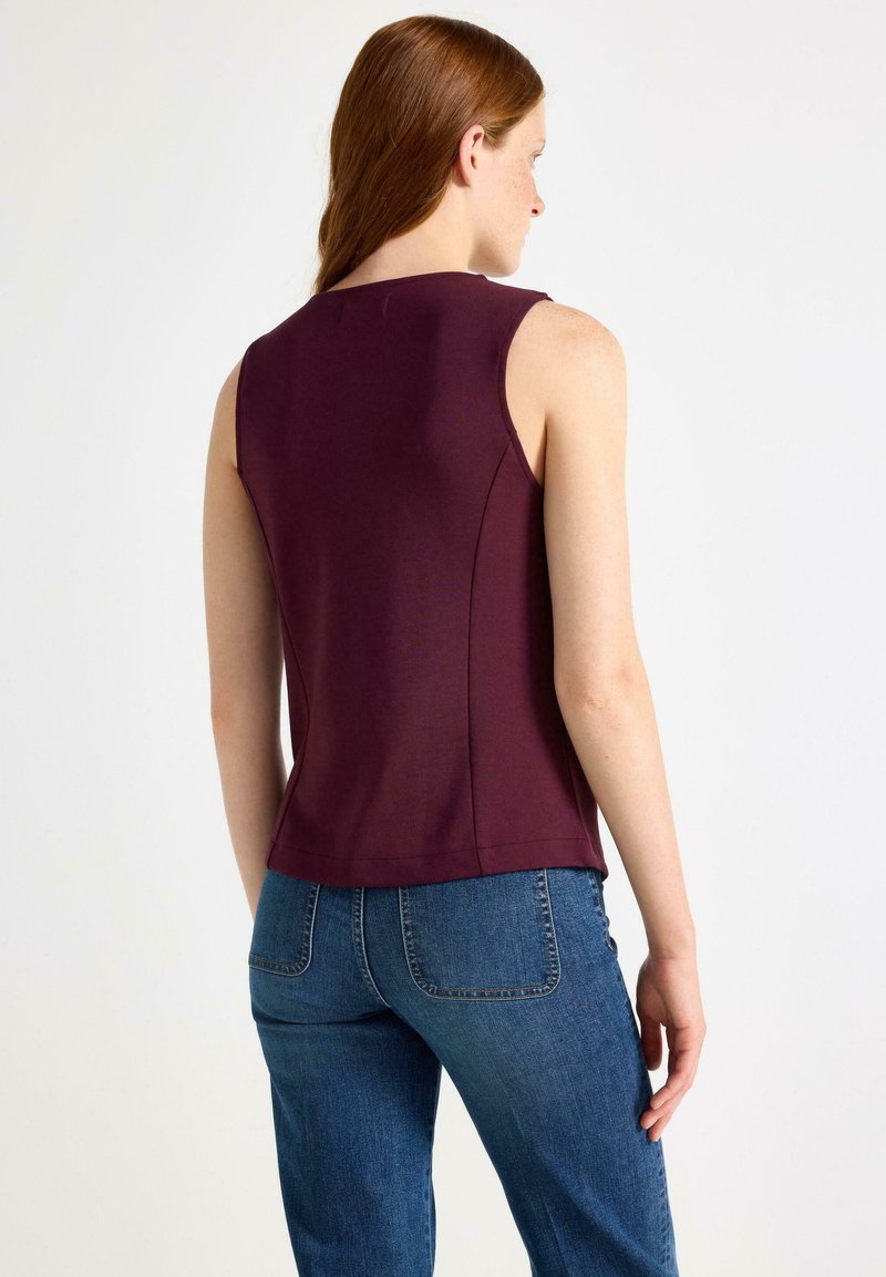 Vrouw met lang rood haar in een mouwloos donkerpaars topje en blauwe jeans, gezien vanaf de achterkant tegen een effen lichte achtergrond.