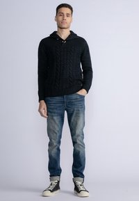 Pulover tricotat de culoare bleumarin, cu model împletit, guler cu glugă și nasturi din lemn, purtat cu blugi albastri decolorati și adidași negri.