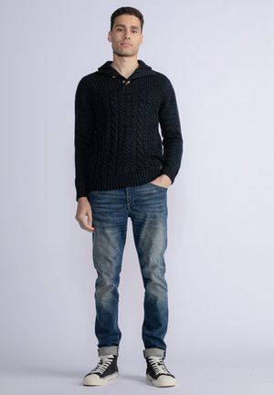 Granatowy sweter dzianinowy z wzorem w warkocze, z kapturem i drewnianymi guzikami, połączony z jasno niebieskimi dżinsami i czarnymi trampkami.