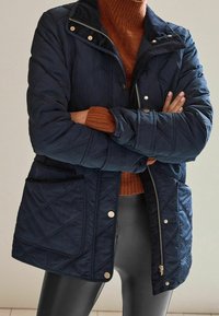 Cappotto trapuntato blu navy con chiusura con zip e bottoni a pressione, caratterizzato da colletto in velluto a coste e due tasche frontali, abbinato a un maglione color ruggine sotto.