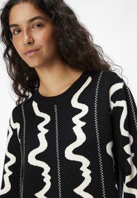 Pull noir avec un motif ondulé blanc, col rond et tissu texturé. Accentué par de fines lignes blanches entre les sections à motifs.