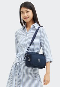 Giovane donna che indossa un vestito a righe verticali blu e bianche, mentre porta una borsa a tracolla blu navy con un charm sulla cerniera.