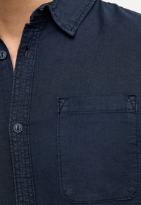 Camisa de botones azul marino oscuro hecha de tela texturizada, con un cuello puntiagudo y un solo bolsillo en el pecho con acentos de costura.